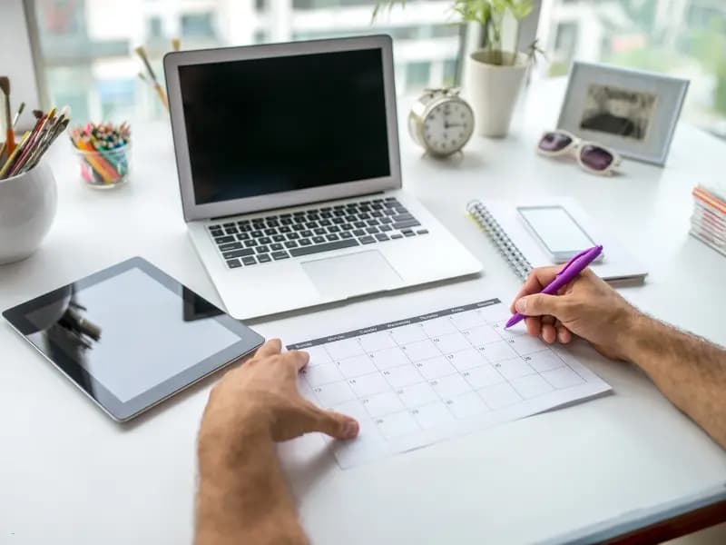 Calendar management with a printed monthly planner, laptop, and tablet on a well-organized desk